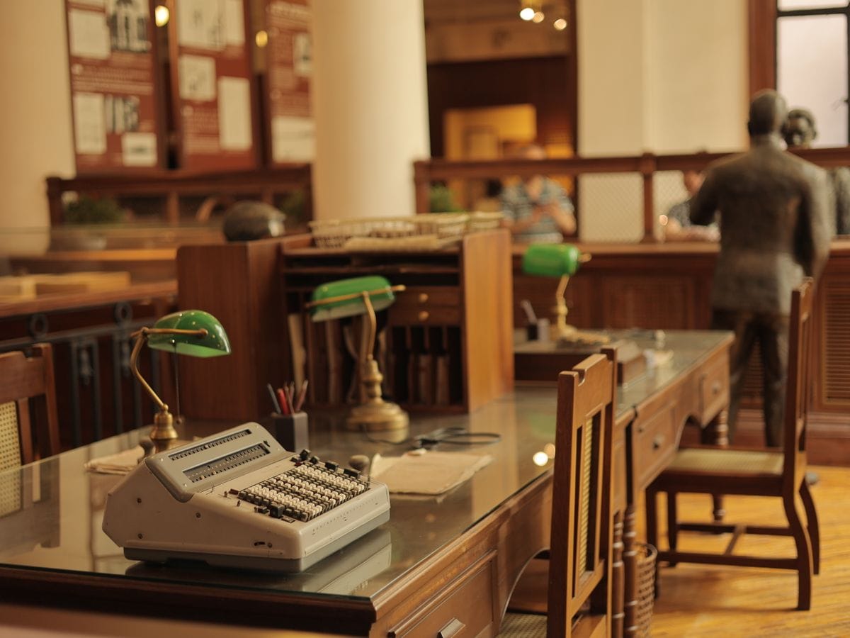 Rows of wooden desks with green banker’s lamps in a records office